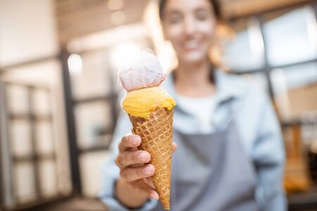 Female cheerful seller holding yummy ice cream in waffle cone indoors, selling sweets in the shopの写真素材