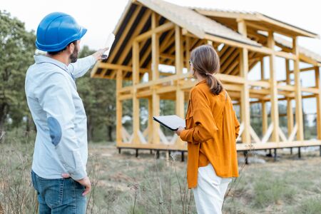 Builder with a female client near the wooden house structure on the construction site outdoors. Building and designing wooden frame house conceptの写真素材