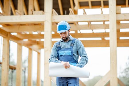 Portrait of a confident builder in blue overalls and hard hat standing with blueprints on the construction site. Building wooden frame house conceptの写真素材