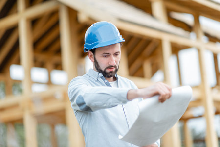 Builder or architect in hard hat supervising a project, standing with blueprints on the construction...