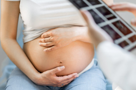 Pregnant woman hugging her belly, sitting on a gynecological chair during a medical examination, cropped view without face focused on abdomenの写真素材