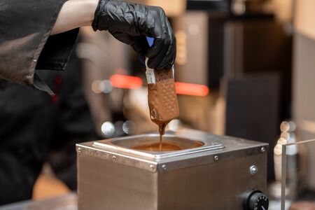 Chef in black workwear dips ice cream into the hot melted chocolate, making ice cream on a stick with toppingsの写真素材