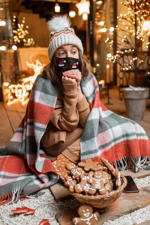 Young woman in facial mask celebrating alone New Year holidays sitting with sweet gingerbreads on a...
