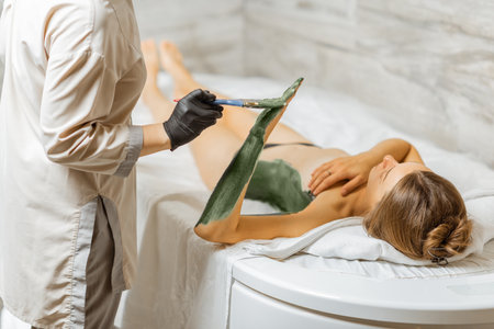 Young woman relaxing during a mud treatment at spa, doctor applies green therapeutic mud on the patients body at the treatment roomの写真素材