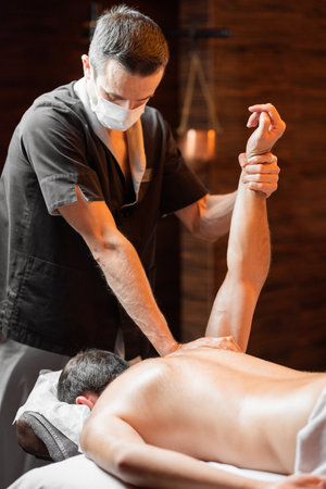Professional masseur in facial mask doing a deep massage to a male client at Spa salon. Business during the epidemic conceptの写真素材