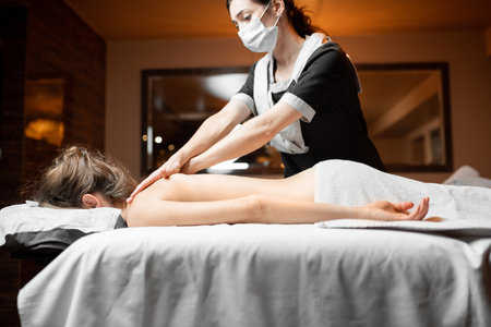 Professional female masseur in facial mask doing massage to a client at Spa salon. Business during the epidemic conceptの写真素材