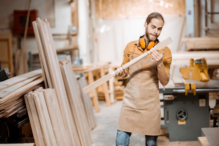 Handsome carpenter in uniform working with wood, checking the quality of the wooden baluster at the joineryの写真素材