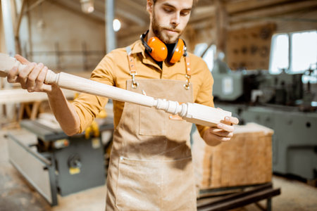 Handsome carpenter in uniform working with wood, checking the quality of the wooden baluster at the joineryの写真素材