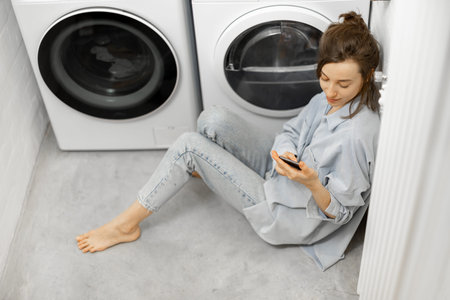 Relaxed woman with a smartphone in the laundry roomの写真素材
