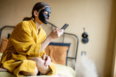 Woman in bathrobe with black cleansing mask and phone in bedroomの写真素材