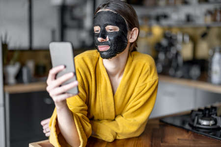 Woman with black cleansing mask on her face on the kitchen at homeの写真素材