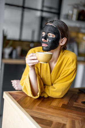 Woman with black cleansing mask on her face on the kitchen at homeの写真素材