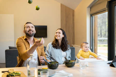Family having fun with their baby son during a lunch time at home. Man juggling lemons. Concept of healthy vegan eating and happy parentingの写真素材