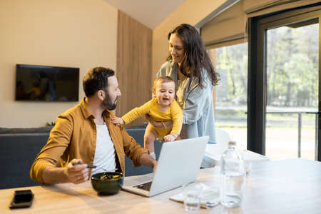 Young father works on a laptop during a lunch time at home with his wife and one year son. Home family routine and work from home conceptの写真素材