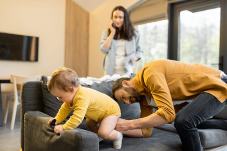Man wearing his baby son on the couch at home. Fatherhood and child care by father concept. Wife...