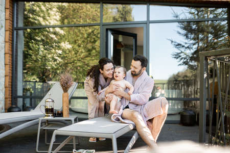 Young family with a one year baby boy resting at Spa resort, sitting on a sunbed near the sauna outdoors. Parents with a newborn son on vacationの写真素材