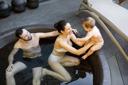 Young family relaxing with a little baby boy at spa, sitting in the hot vat outdoors. Family on...