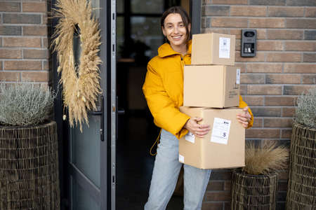 Housewife receiving goods purchased online on the porch of her house, holding cardboard boxes at the entrance door. Concept of contactless food delivery home during a pandemicの写真素材