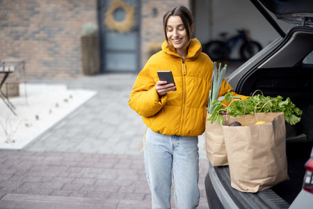 Woman arrives home with a groceries, standing with phone near the car trunk. Shopping healthy food in eco-packaging and lifestyle conceptの写真素材