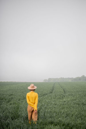 Woman in hat walking on green wheat field during foggy weather