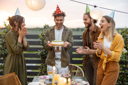 Young adult friends celebrating birthday, having fun blowing candles on a cake at backyard of a country house outdoorsの写真素材