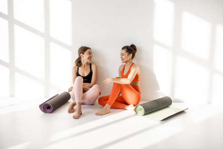 Two female friends in sportswear sitting together with yoga mats and having talk during a yoga training break at gym on the white backgroundの写真素材