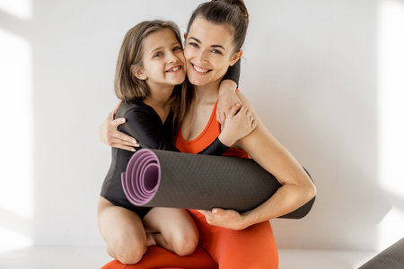 Portrait of a cheerful sports mom and little daughter sitting together with yoga mats during sports activities indoors. Close relationships with mother and sports in childhoodの写真素材
