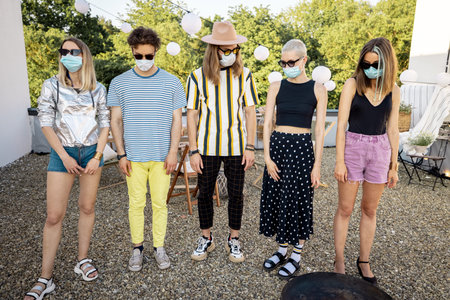 Sad group of stylish friends in face masks standing in a row outdoors. Gathering for a party on a...