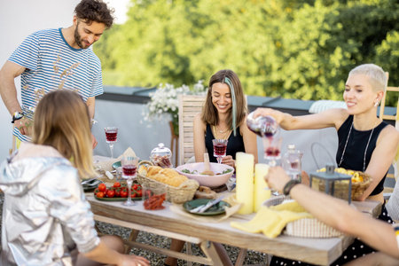 Young group of stylish people having a festive dinner on the roof terrace. Hipsters drinking wine...