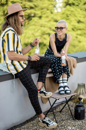 Two stylish carefree persons talk and drink beer at roof top terrace at countryside house. Friends...
