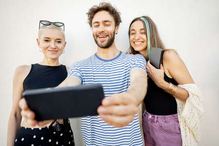 Young stylish friends have fun, taking selfie photo on phone on a white background. Millennials or gen z spend time together with digital gadgetsの写真素材
