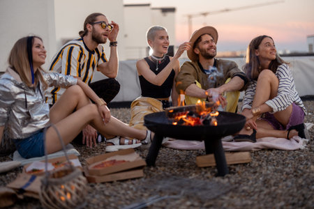 Group of young stylish friends sitting together by the fireplace, having a picnic on the rooftop...