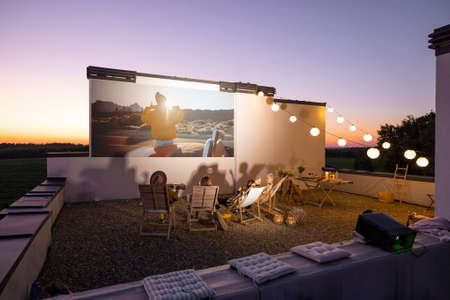 Small group of people watching movie on the rooftop terrace at sunset. Open air cinema concept. Romantic leisure and entertainment on the roof of a country houseの写真素材