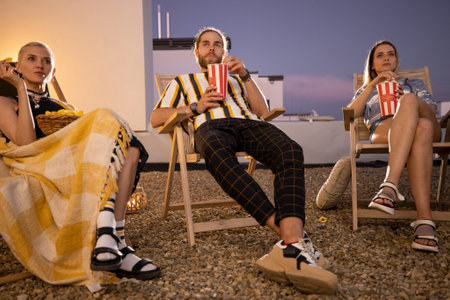 Young people watching football match or some film with popcorn outdoors on a rooftop terrace at...