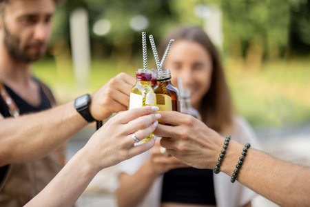 People clinking bottles with a strong alcohol drinks on a picnic outdoors, close-up. Holding bottles...