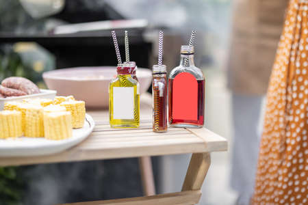 Bottles with liqueur or berry tinctures on a table with food for bbq, people grilling on a background, close-up on bottles with blank labels for copy pasteの写真素材