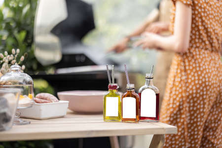 Bottles with liqueur or berry tinctures on a table with food for bbq, people grilling on a background, close-up on bottles with blank labels for copy pasteの写真素材