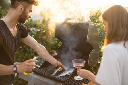 Men cooking fresh trout fish on a modern gas grill outdoors at sunset. Cooking healthy food on the...