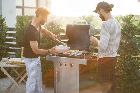 Two caucasian guys cooking vegetables and fish on a modern gas grill at backyard on a sunset. Healthy eating on the open air, male friendshipの写真素材