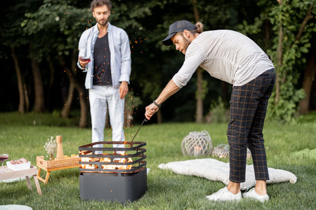 Two guys light a fire in a barbecue preparing for a picnic on a green lawn near the forest in the...
