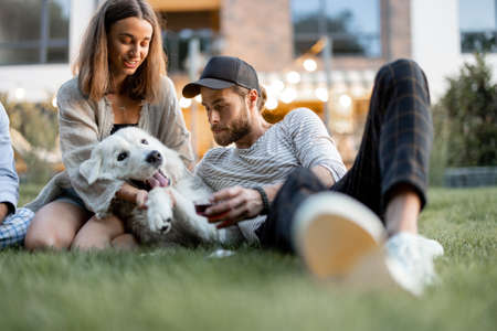 Young couple having fun playing with a dog and drinking wine, sitting on the green lawn at backyard of the country house in the eveningの写真素材