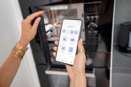 Smart phone with running mobile application to control smart devices in the kitchen. Female controlling microwave or oven with phone at home. Smart home conceptの写真素材