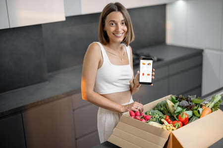 Woman buying fresh vegetables online, standing with package full of food and holding smart phone with running e-shop application. Buying online and delivering groceries conceptの写真素材