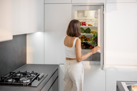 Young woman standing near the fridge full of fresh vegetables at modern kitchen. Healthy vegan eating conceptの写真素材