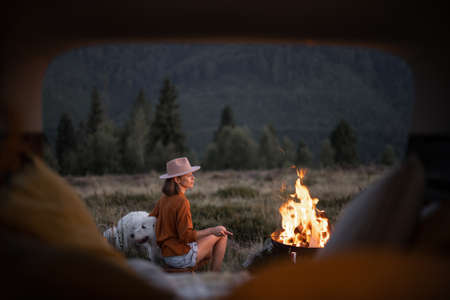 Woman sitting with dog near bonfire at picnic in the mountains, inside view from a car trunk. Camping and vacation in the mountains, travel by car conceptの写真素材