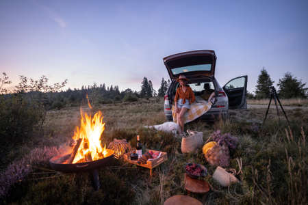 Woman enjoys beautiful view on the mountains, having a picnic with fireplace and sitting in the vehicle trunk at dusk on the evening. Traveling by car in natureの写真素材