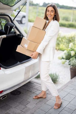Portrait of a young business woman picking up parcels from a car trunk, coming home by car. Concept of buying goods online and delivering them homeの写真素材