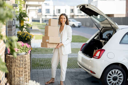 Young business woman picking up parcels from a car trunk, coming home by car. Concept of buying goods online and delivering them homeの写真素材