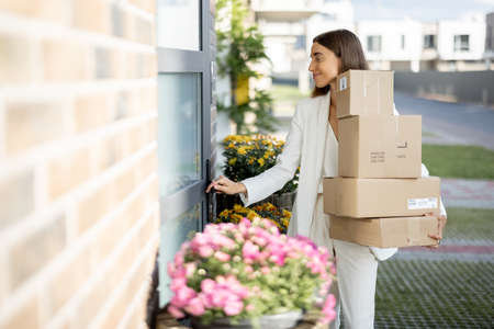 Young woman carrying parcels home, standing with a heap of cardboard boxes on the porch of her house. Concept of buying goods online and delivering them homeの写真素材