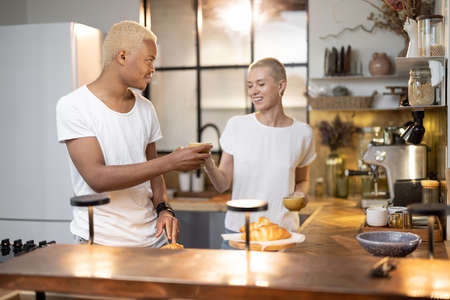 Multiracial couple drinking and toasting drink at home kitchen. Concept of relationship. Idea of modern domestic lifestyle. Black man and european girl spending time together in modern flatの写真素材